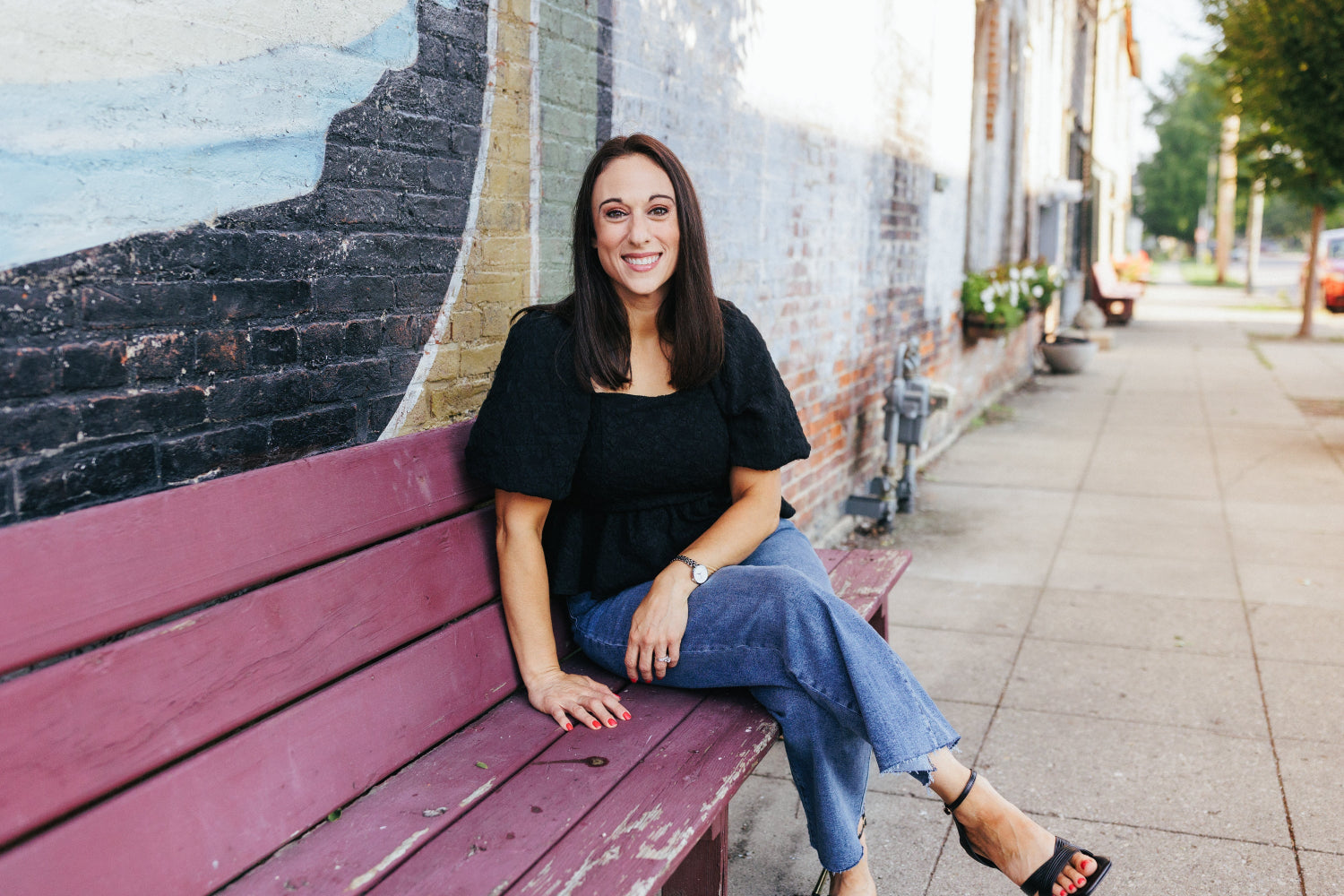 Stylish woman in black puff sleeve blouse, and jeans sitting on bench by mural wall