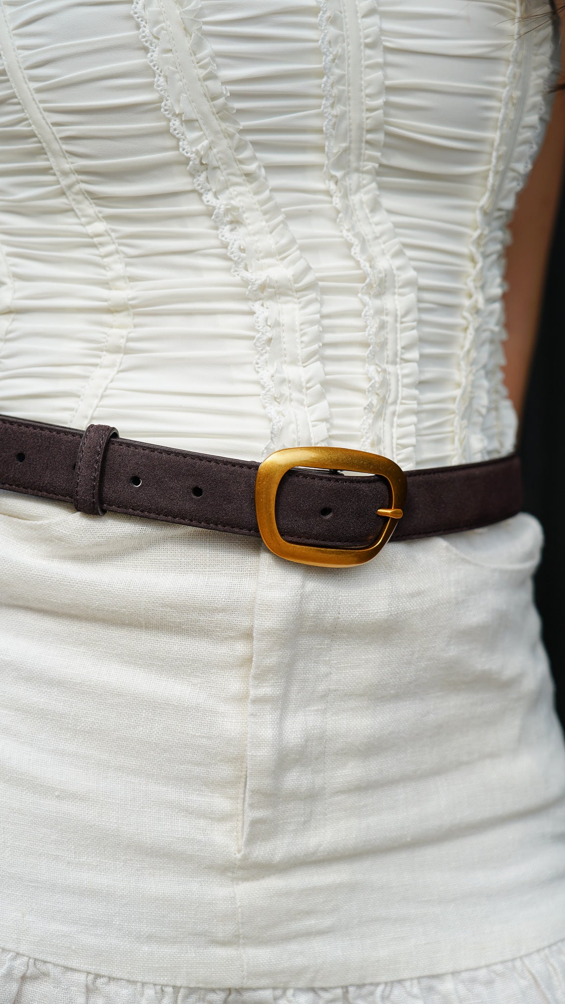Close-up of a white textured dress with an elegant brown suede waist belt with curved gold buckle