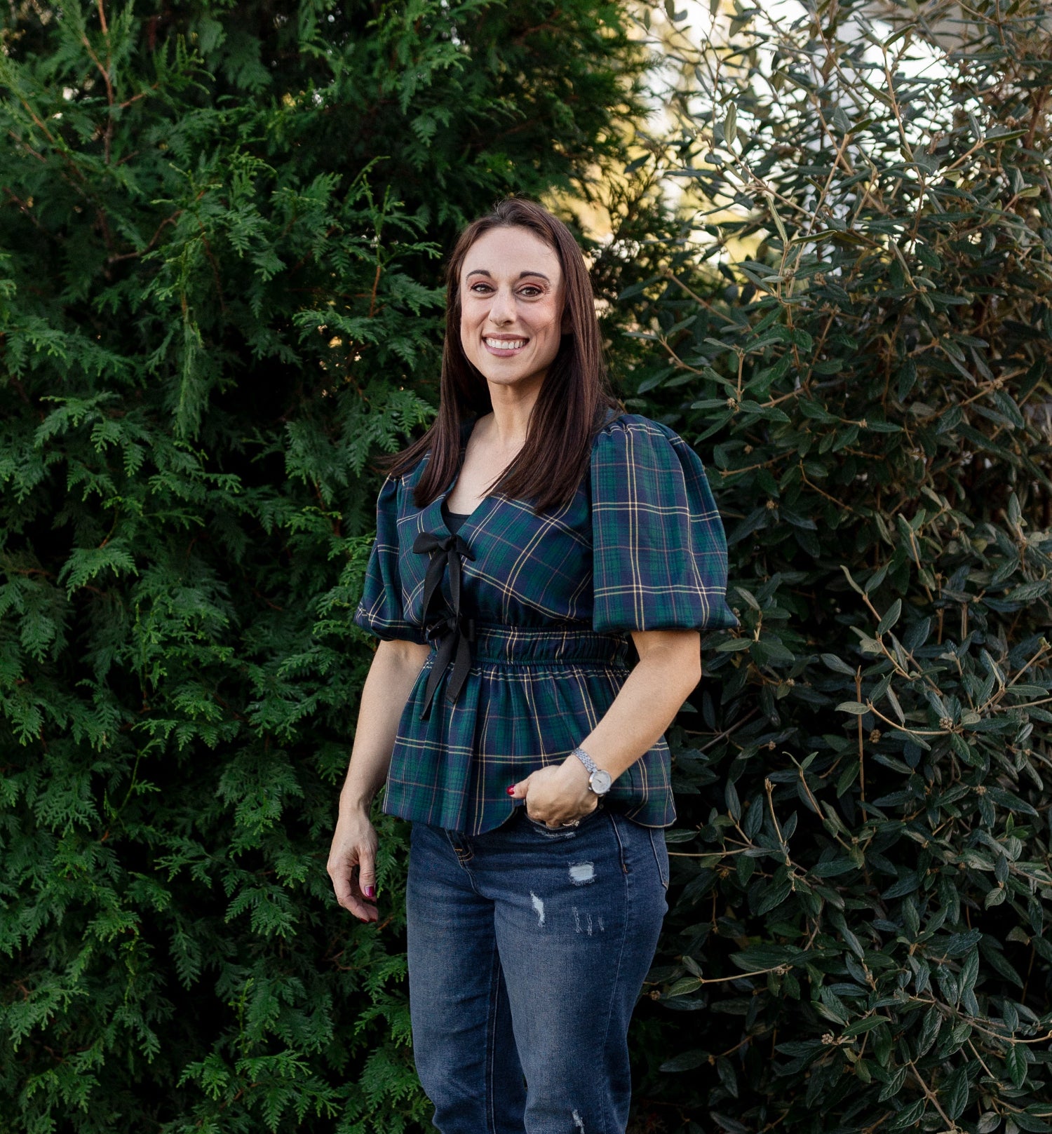 Woman wearing a plaid shirt and jeans standing in front of green bushes