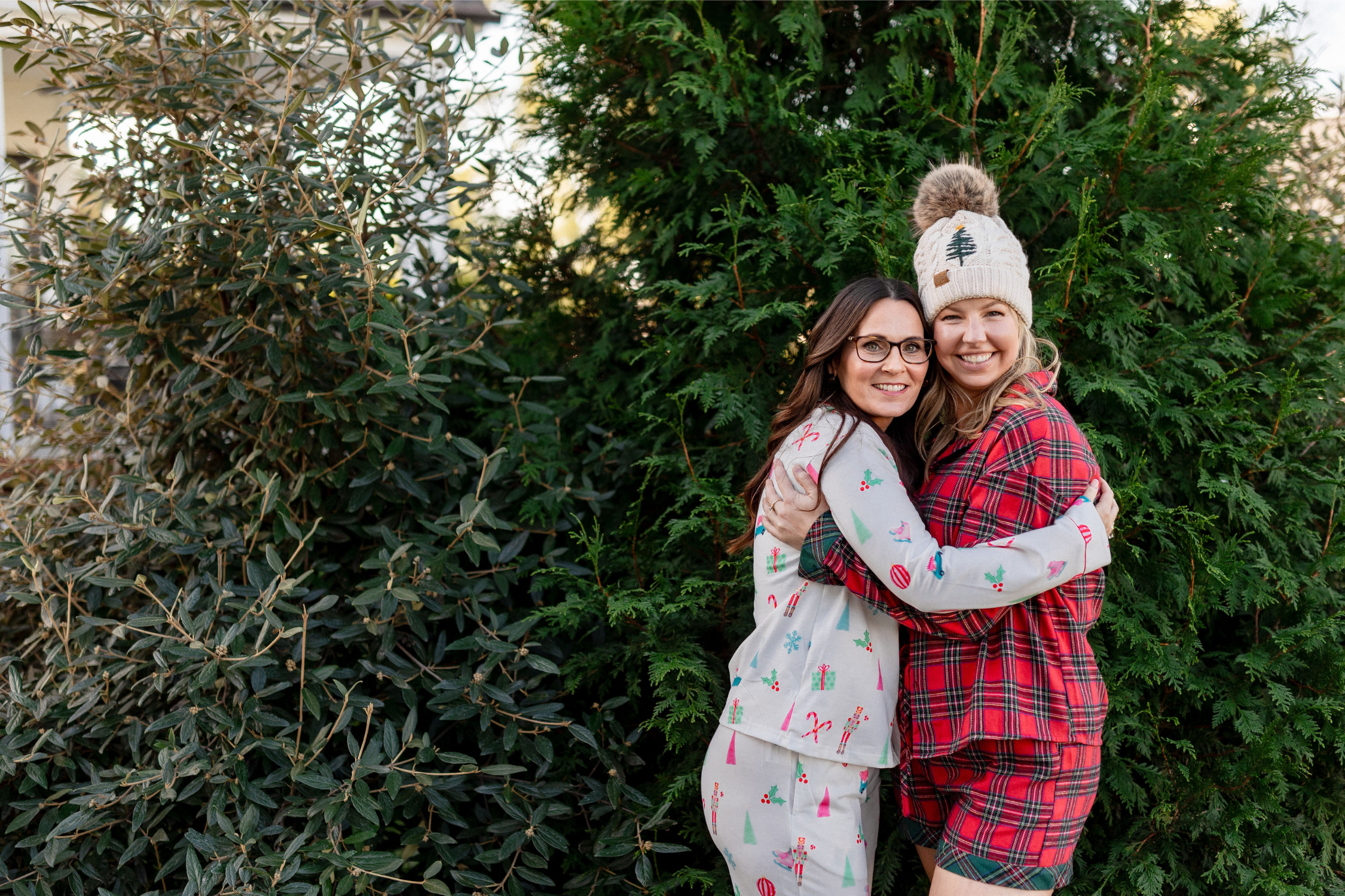 Two women smiling and hugging outdoors in cozy Christmas pajamas, standing in front of evergreen trees