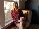 Woman in a red plaid 2 piece pajama set sitting by a window holding a mug.