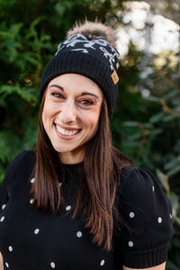 Woman wearing a black knit beanie with a pom-pom and a black sweater with white polka dots, standing outdoors.