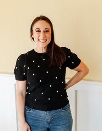Woman wearing a black polka dot sweater and blue jeans standing against a beige wall.