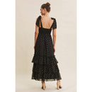 Back view of woman wearing a black polka dot dress with a tiered skirt on a beige background