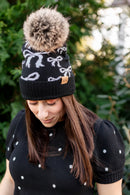 Woman wearing a black beanie with white bow pattern and a fluffy pom-pom, standing outdoors.