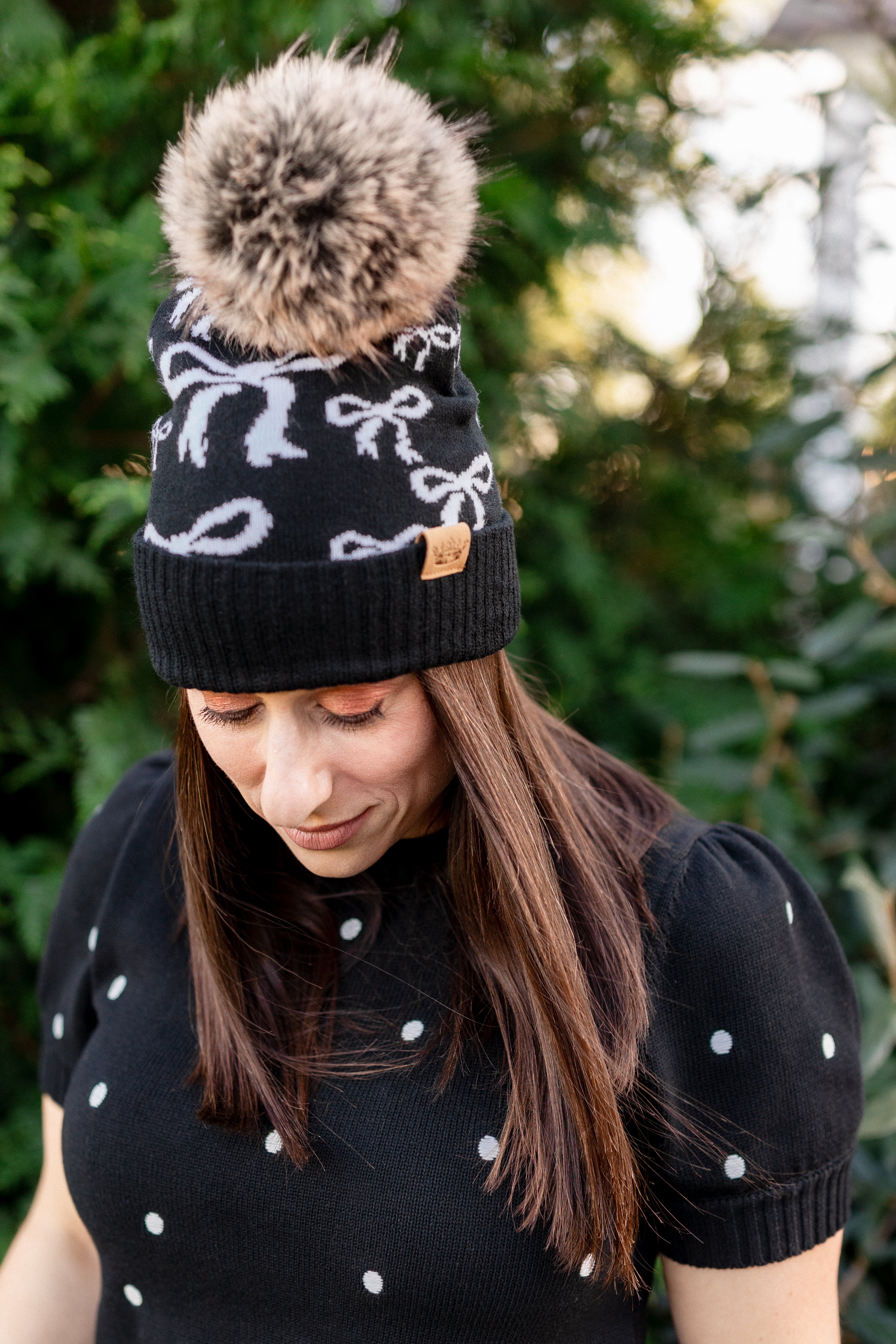Woman wearing a black beanie with white bow pattern and a fluffy pom-pom, standing outdoors.