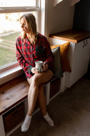 Woman sitting by a window holding a mug, wearing a 2 piece plaid pajamas set and white socks.