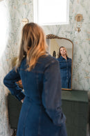 Woman in a blue denim dress standing in front of a mirror with floral wallpaper.