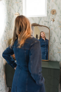 Woman in a blue denim dress standing in front of a mirror with floral wallpaper.