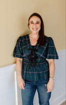 woman wearing green tartan top and dark wash jeans standing against a neutral wall.