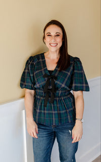 woman wearing green tartan top and dark wash jeans standing against a neutral wall.