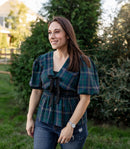 Woman wearing a plaid shirt and jeans standing outdoors with trees and a fence in the background