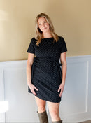 Woman in a denim polka dot side tie dress standing in a room with beige walls and white baseboards.