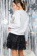 Back view of woman wearing a light gray sweater and black sequin skirt against a silver tinsel background.